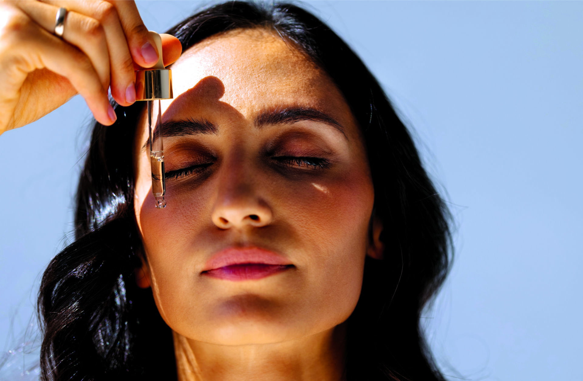 Close-up of charming woman with her eyes closed holding pipette with regenerating serum with hyaluronic acid. Attractive girl with beautiful healthy skin and moisturizing anti-aging facial essence.