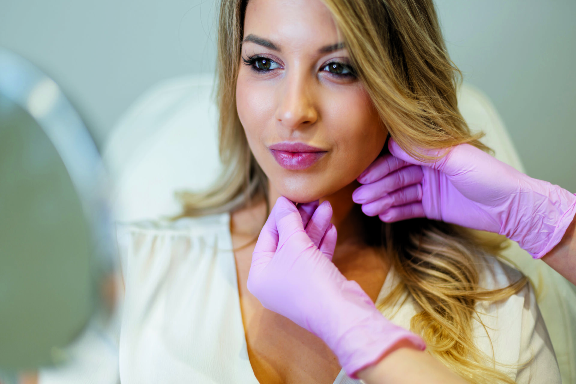 Piękna, świeża kobieta o czystej skórze twarzy i ust w gabinecie kosmetycznym. Profesjonalna kosmetyczka w różowych rękawiczkach wykonująca zabiegi.
