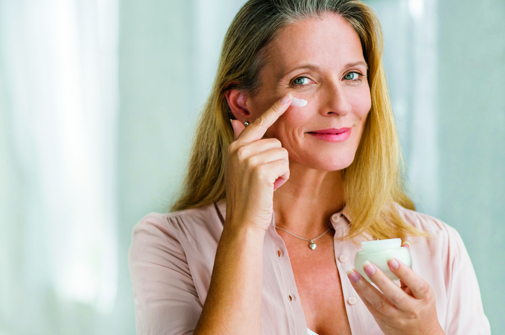 Uśmiechnięta starsza kobieta nakłada krem przeciwstarzeniowy, aby usunąć cienie pod oczami. Szczęśliwa dojrzała kobieta używa kremu kosmetycznego, aby ukryć zmarszczki pod oczami. Kobieta używa kremu nawilżającego na dzień, aby przeciwdziałać starzeniu się skóry.