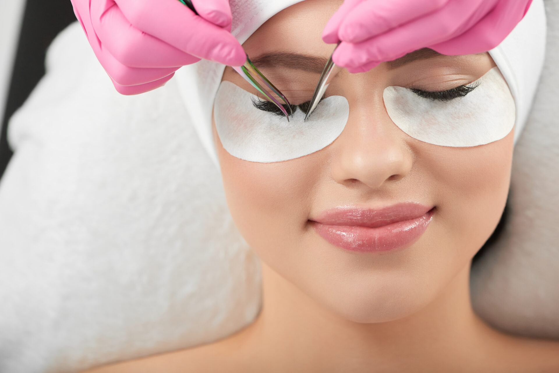 Profesjonalne zdjęcie uśmiechniętej modelki leżącej podczas zabiegu przedłużania rzęs. Przedłużanie rzęs klientce w różowych rękawiczkach higienicznych. Mistrzyni używa różnych rodzajów pęset. Dziewczyna z zamkniętymi oczami.