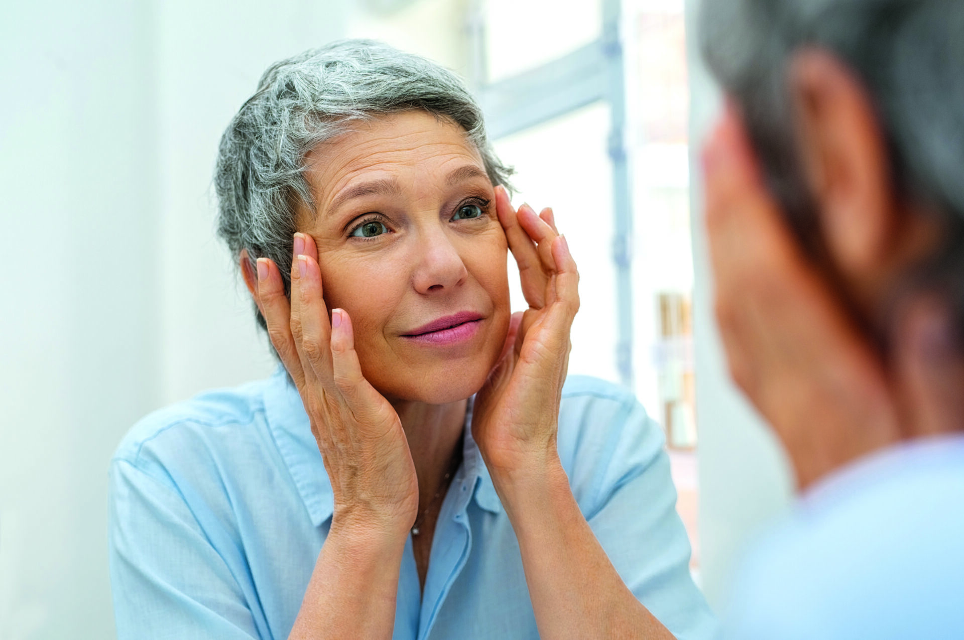 Piękna starsza kobieta sprawdzająca stan skóry twarzy i szukająca niedoskonałości. Portret dojrzałej kobiety masującej twarz i przyglądającej się zmarszczkom wokół oczu w lustrze. Starsza kobieta o siwych włosach sprawdzająca zmarszczki wokół oczu, proces starzenia się.