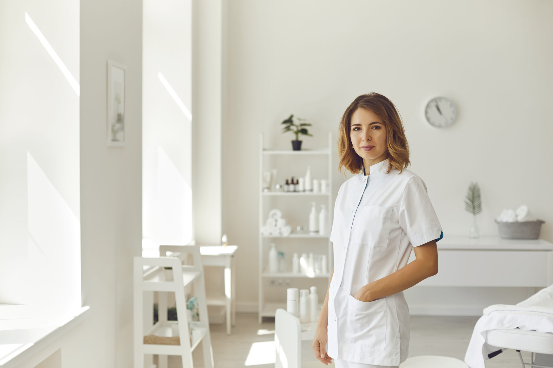 Młoda, uśmiechnięta kosmetyczka lub dermatolog stojąca i patrząca w obiektyw aparatu w jasnym salonie kosmetycznym, na tle kosmetyków. Koncepcja kosmetyki i pielęgnacji skóry