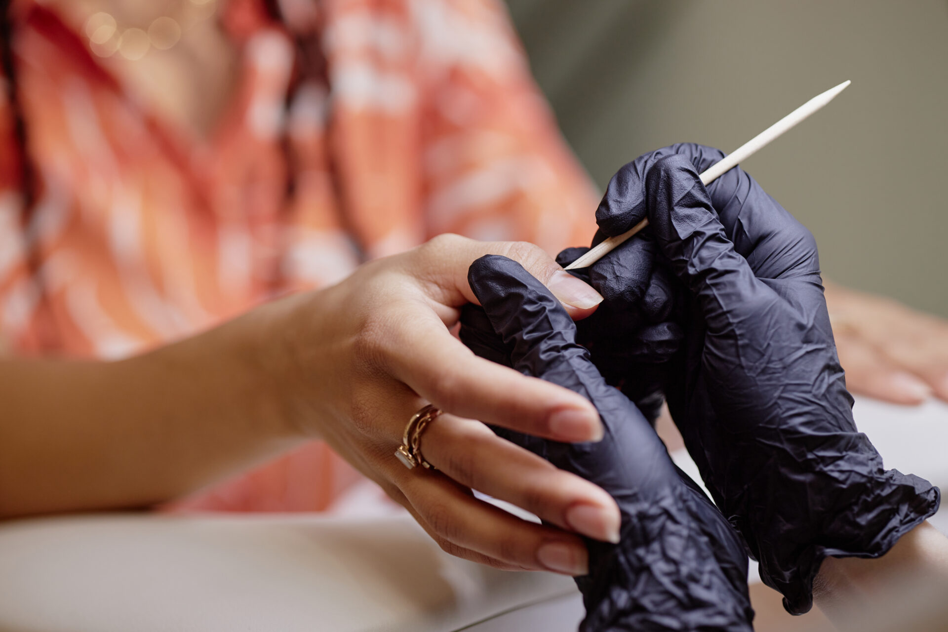 Młoda kobieta mieszanego pochodzenia etnicznego podczas manicure wykonywanego przez profesjonalistkę w czarnych rękawiczkach; widać jej dłonie, gdy kosmetyczka używa drewnianego patyczka do skórek podczas zabiegu pielęgnacji paznokci