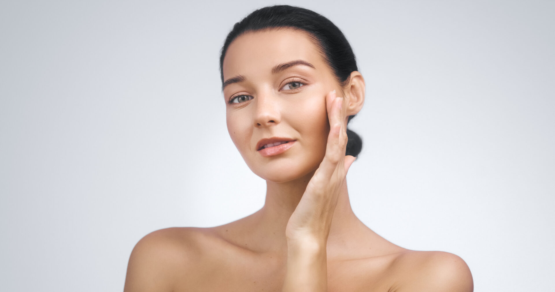 Modelka o promiennym wyglądzie i świetlistej cerze, dotykająca policzka – koncepcja portretu kosmetycznego. Profesjonalna fotografia studyjna, idealna do reklam zabiegów pielęgnacyjnych i kampanii promocyjnych produktów kosmetycznych.