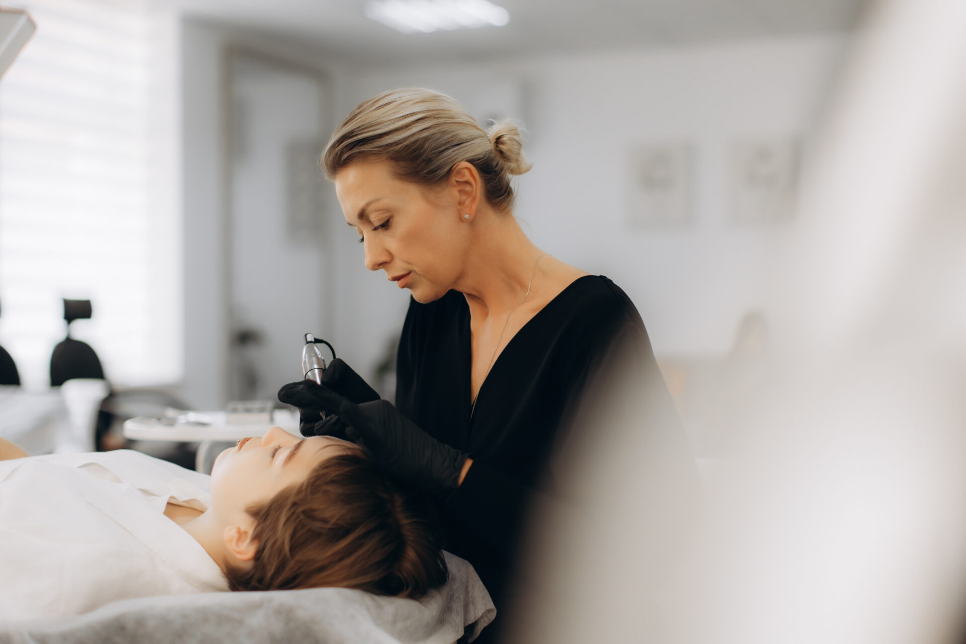 Profesjonalna kosmetyczka wykonująca makijaż permanentny metodą mikrobladingu na brwiach młodej kobiety w nowoczesnym salonie kosmetycznym