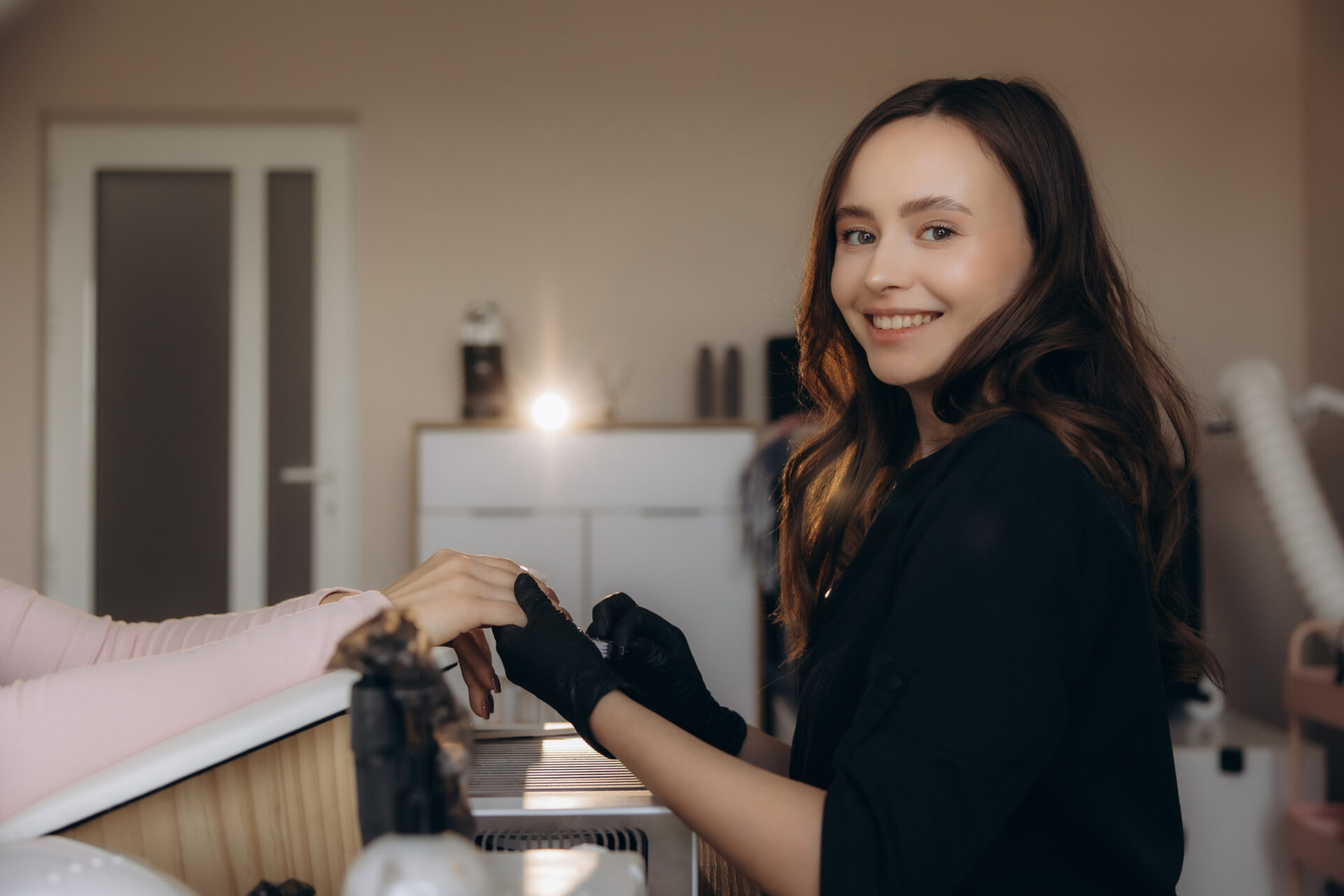Uśmiechnięta stylistka paznokci w rękawiczkach, zajmująca się paznokciami klientki, oferująca zabiegi kosmetyczne i pielęgnacyjne w salonie