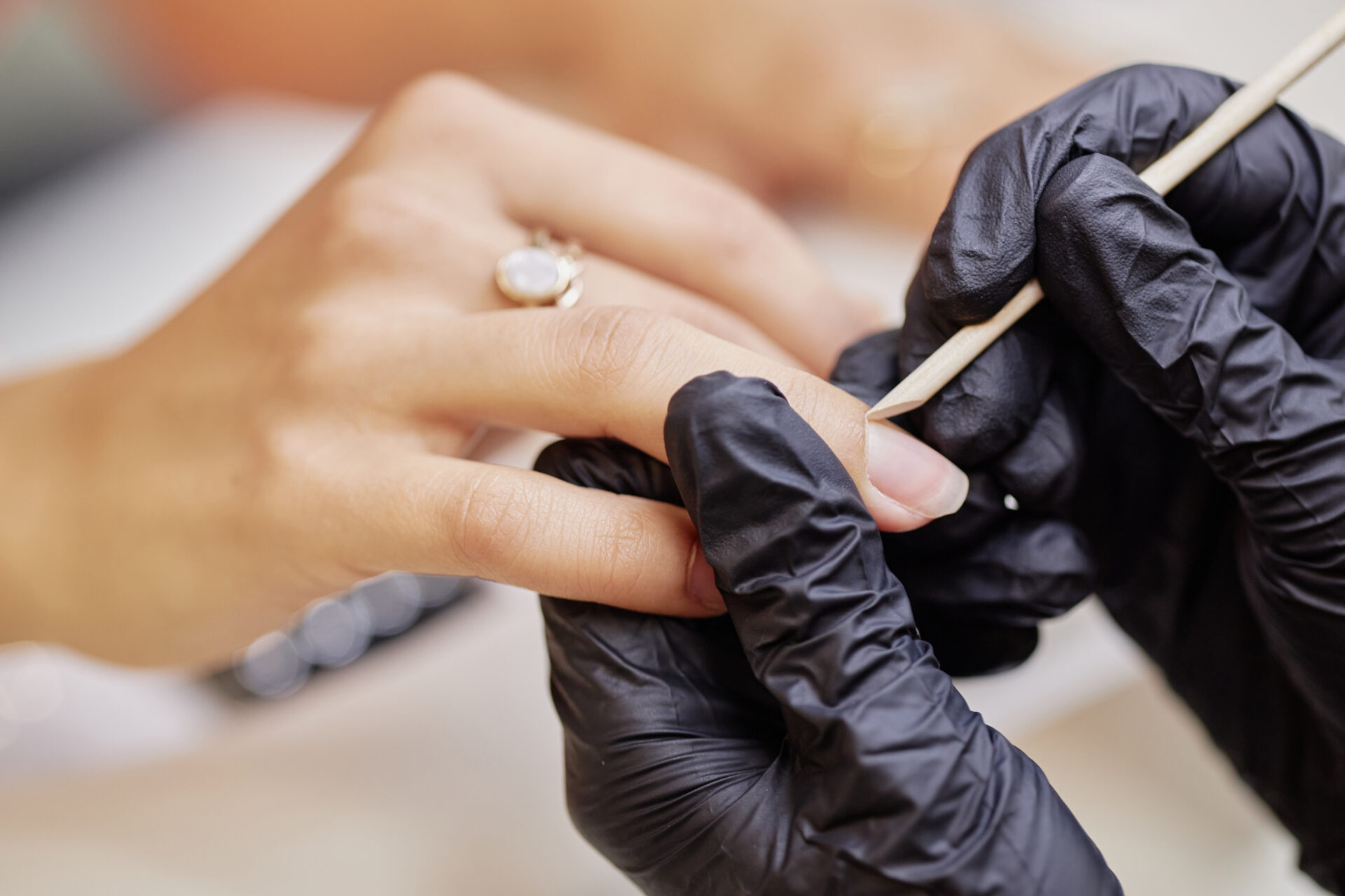 Młoda kobieta podczas manicure wykonywanego przez profesjonalistkę w czarnych rękawiczkach; widać jej dłonie, gdy kosmetyczka modeluje paznokcie drewnianym patyczkiem w ramach zabiegu pielęgnacyjnego