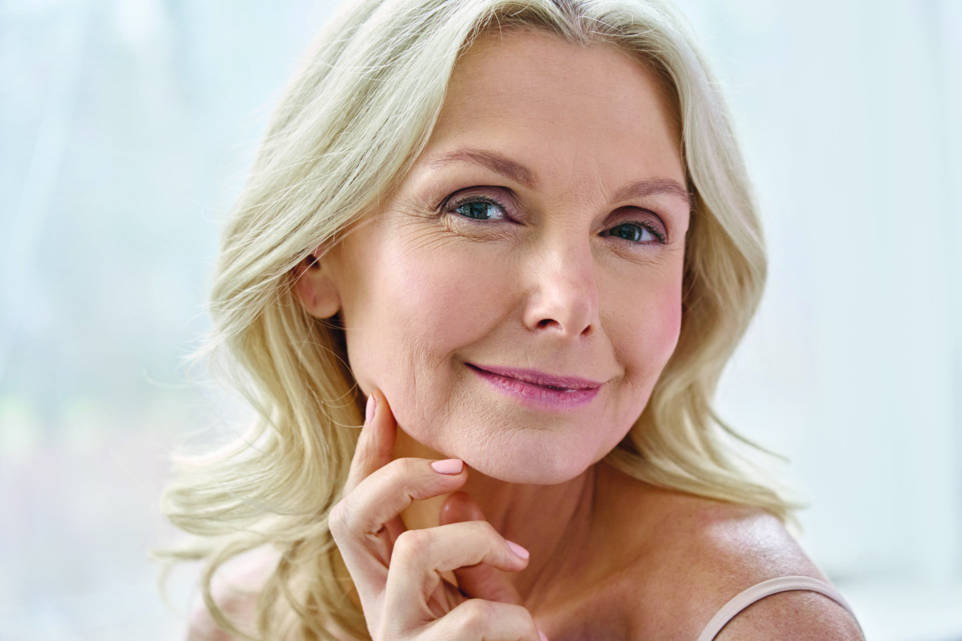 Uśmiechnięta, szczęśliwa i atrakcyjna blondynka w wieku około 50 lat, kobieta w średnim wieku, starsza pani patrząca w obiektyw aparatu, reklamująca kosmetyki przeciwstarzeniowe do pielęgnacji twarzy i ciała, pozująca w łazience. Portret z bliska, ujęcie twarzy