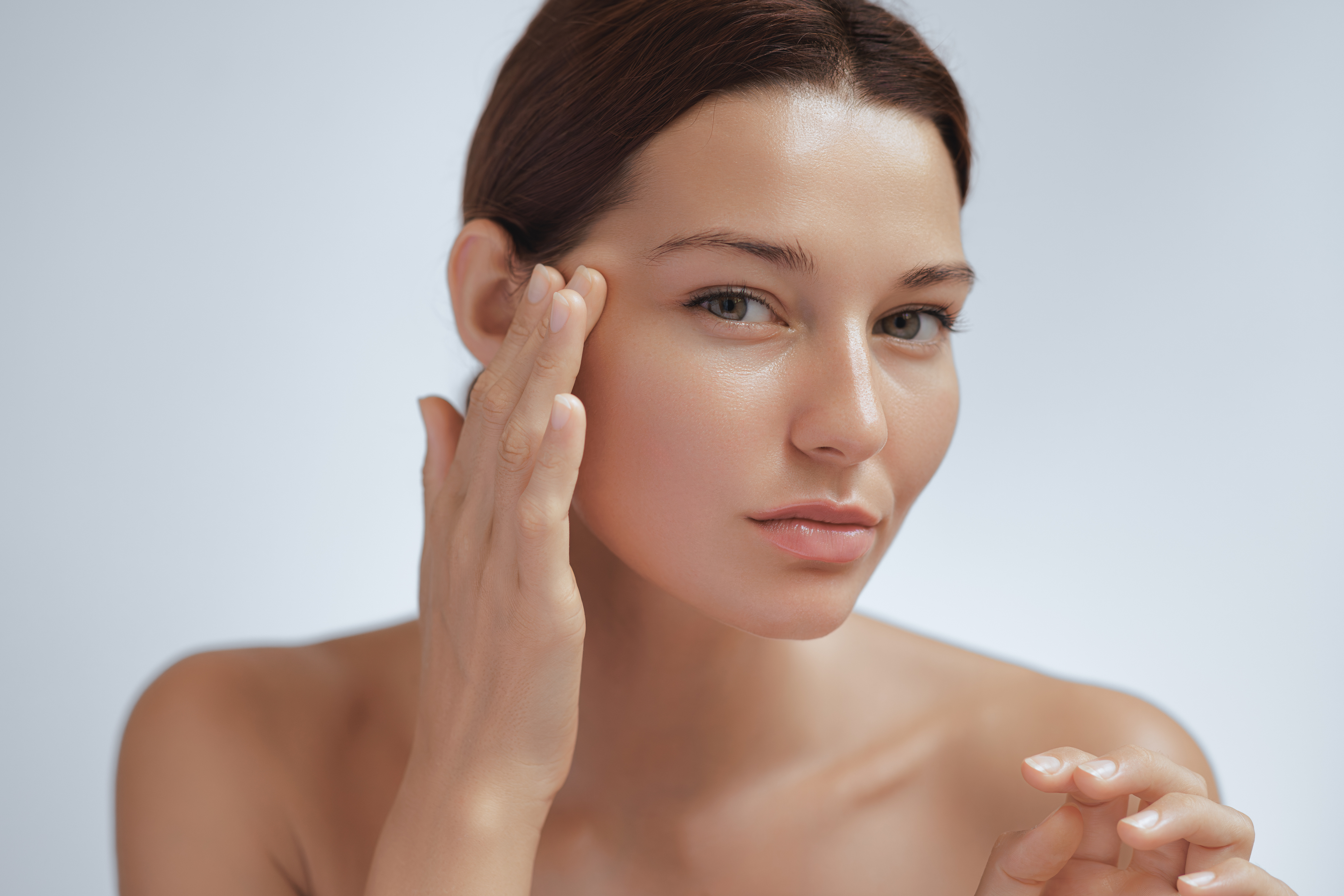 Unhappy young woman touching her eye area and looking at her skin with concern. Ideal image for skin aging, under-eye skincare, cosmetic dissatisfaction, and facial care promo materials.