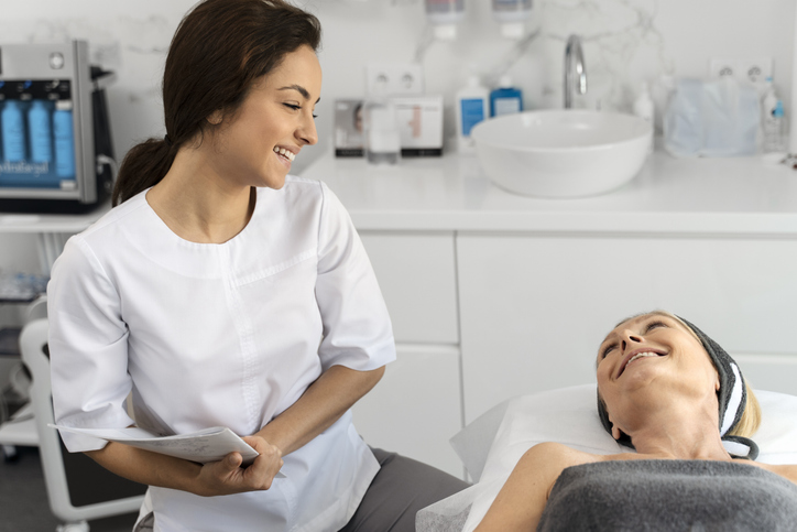 Portrait of charming elderly woman laying on daybed and looking at beautician with smile while she talking with her and holding folder