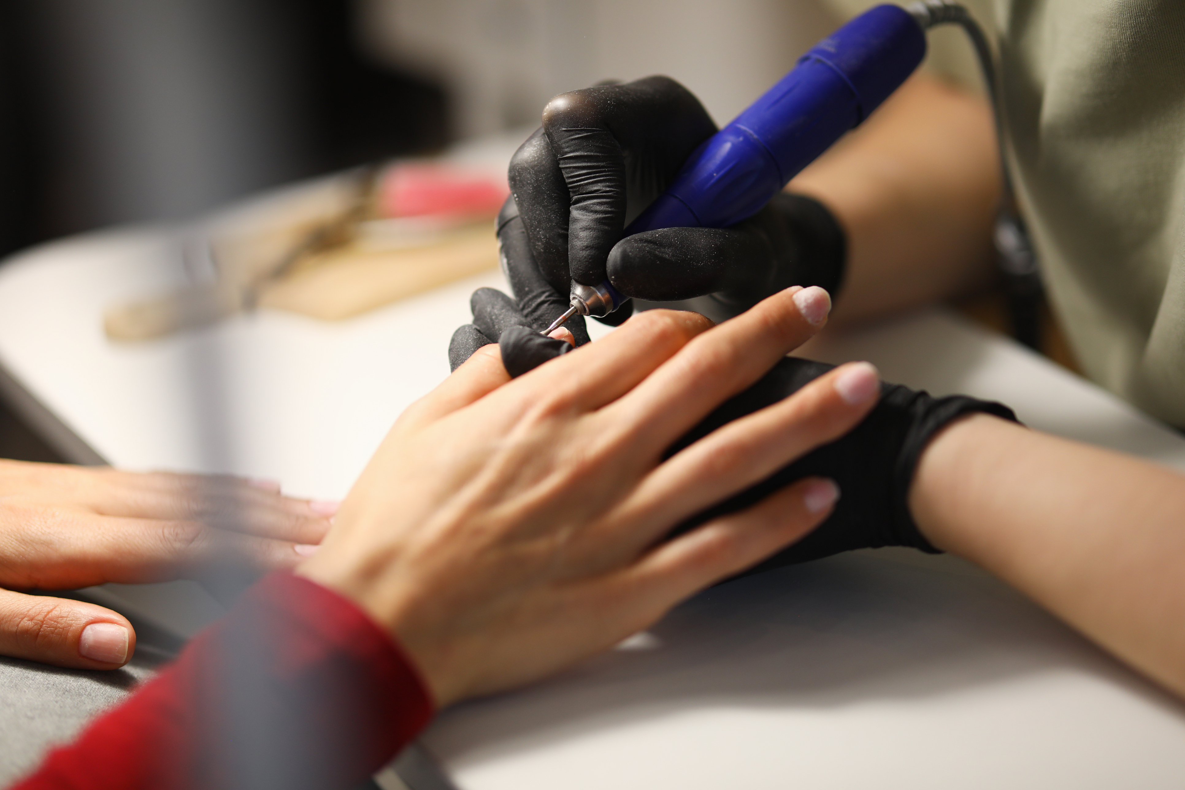 Specjalistka w czarnych rękawiczkach wykonuje zmechanizowany manicure klientce w salonie. Kobieta przygotowuje paznokcie przed nałożeniem lakieru na stanowisku pracy.