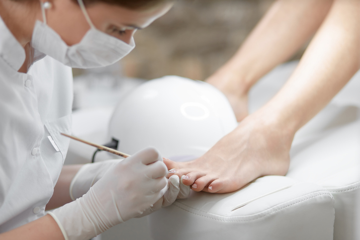 Zbliżenie na profesjonalne oczyszczanie skórek wokół paznokci stóp za pomocą drewnianego patyczka. Specjalistka w masce na twarzy wykonująca pedicure klientce, obok znajduje się lampa UV. Salon kosmetyczny i usługi.